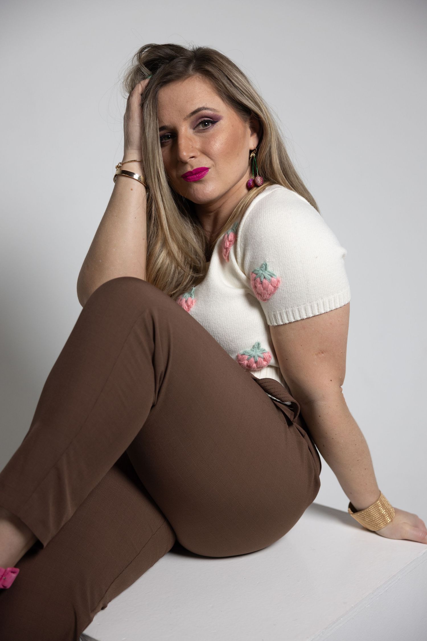 Woman in white strawberry knit top and brown pants sitting relaxed on a white surface in a studio.