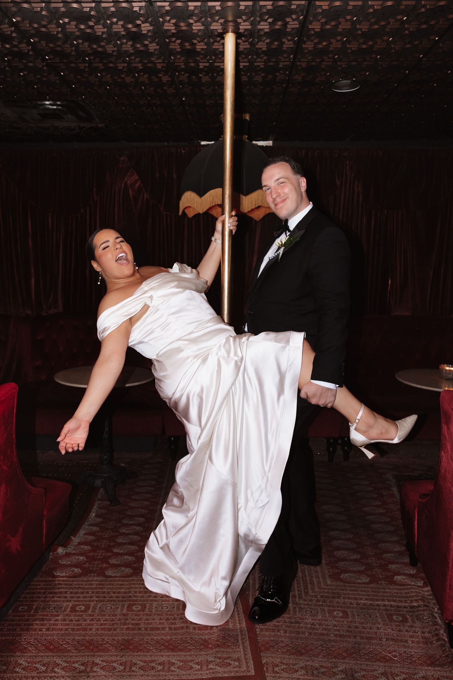 Playful bride and groom pose with a gold pole in a dark, elegant reception venue.