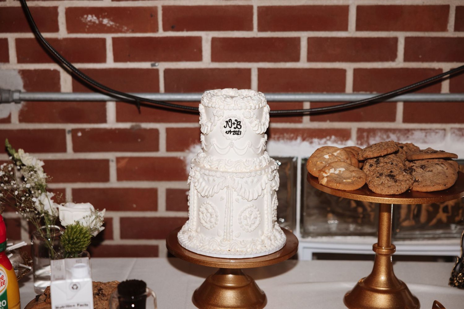 Two-tier white wedding cake with decorative frosting displayed on a gold stand near a brick wall.