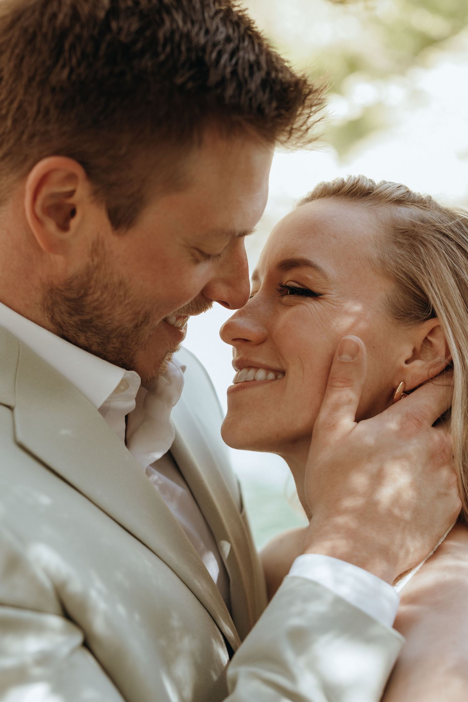 Close-up of smiling bride and groom about to kiss outdoors in soft dappled sunlight.