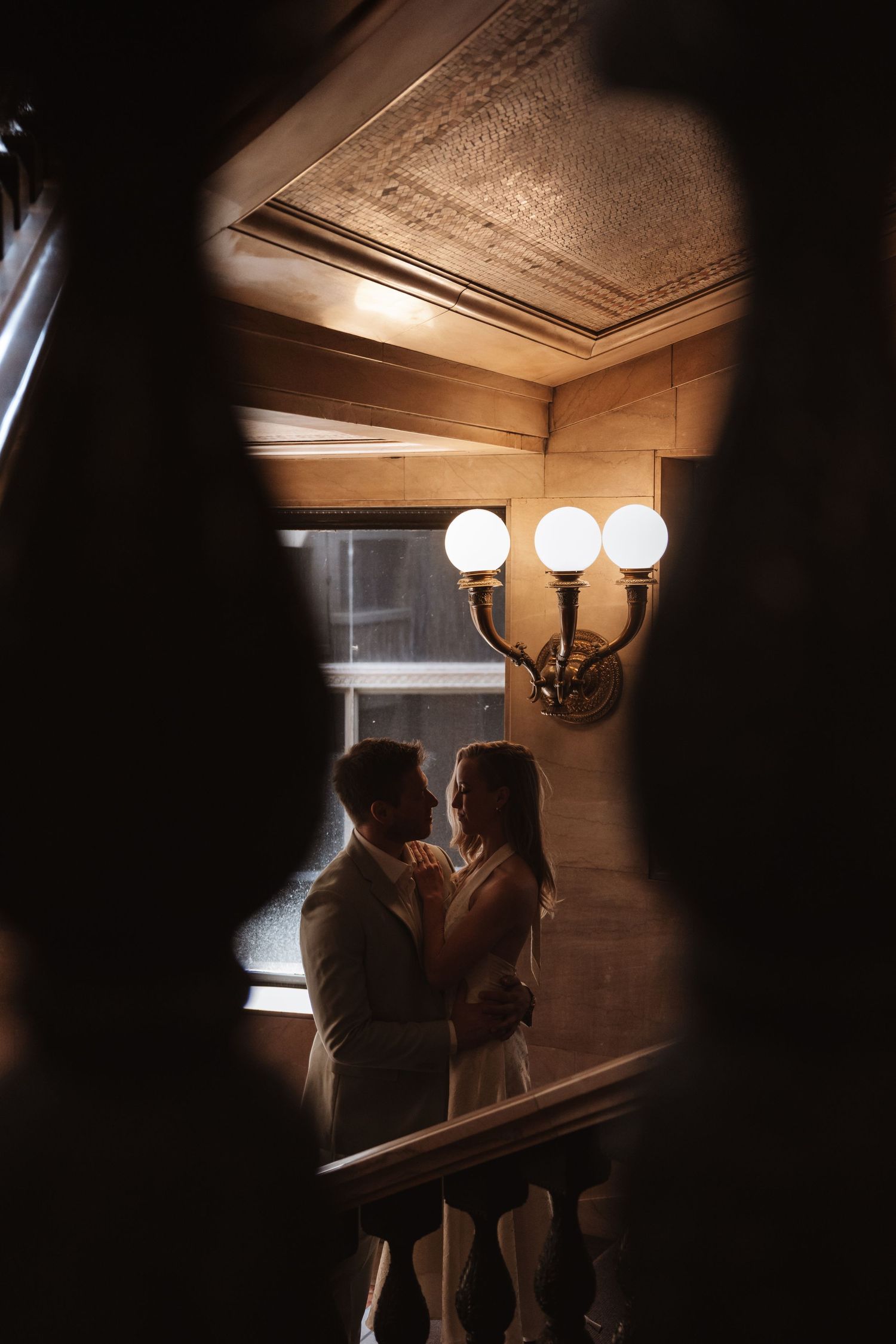 Wedding couple embracing tenderly in warmly lit interior hallway with vintage wall sconce.