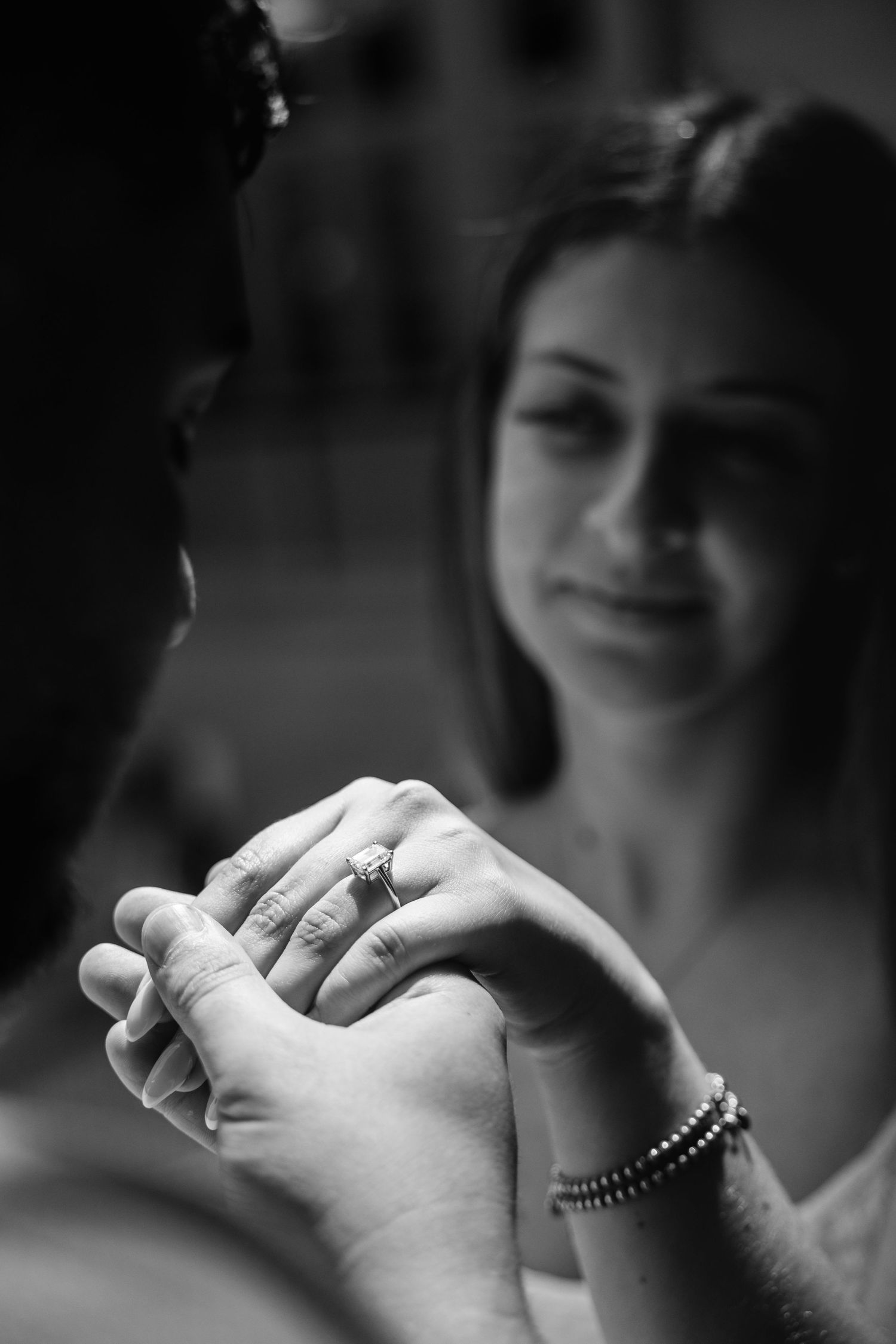 Black and white close-up of couple holding hands showing engagement ring, woman gazing softly in background.