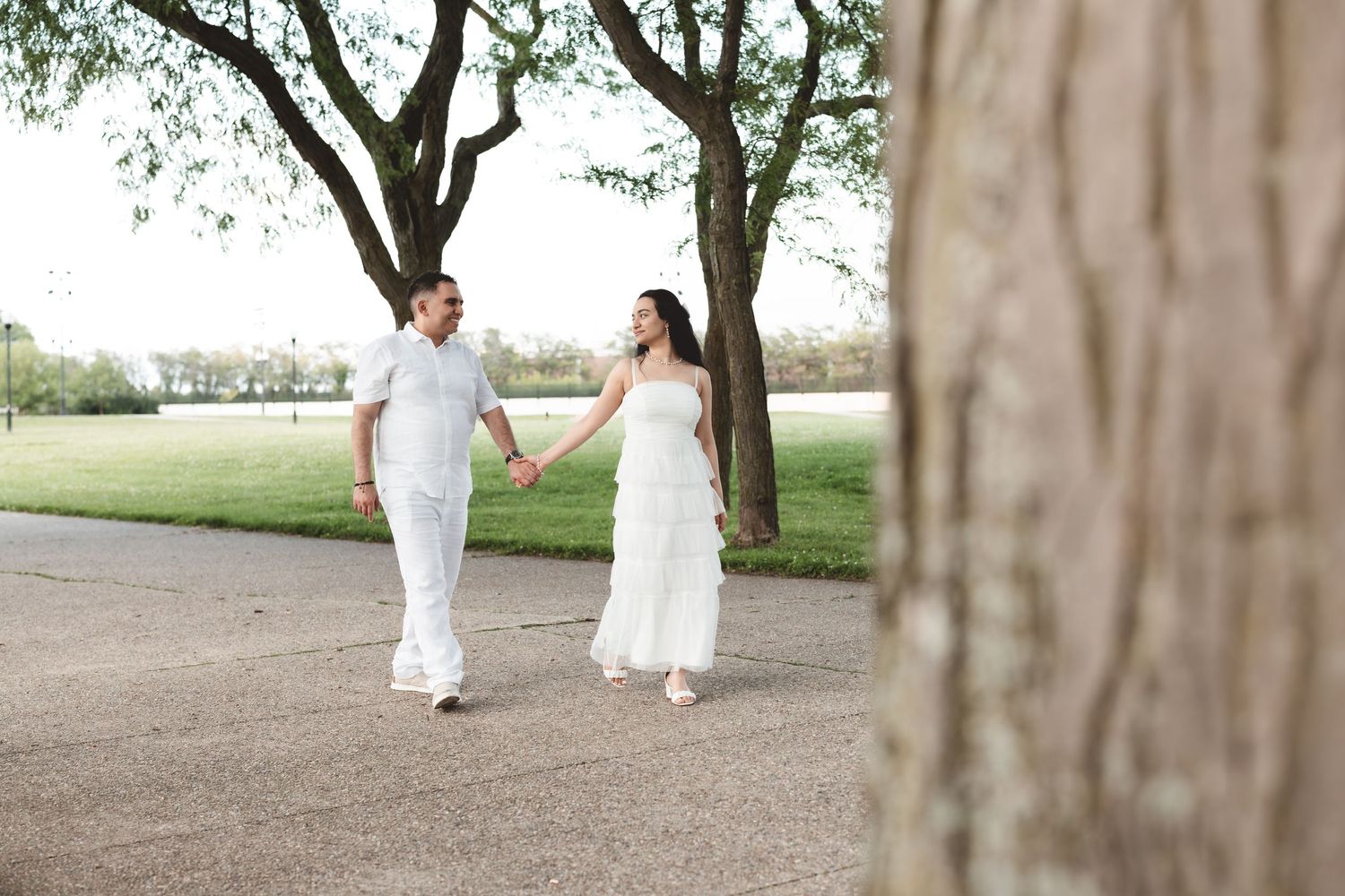 Couple in white outfits holds hands walking along tree-lined path in park.