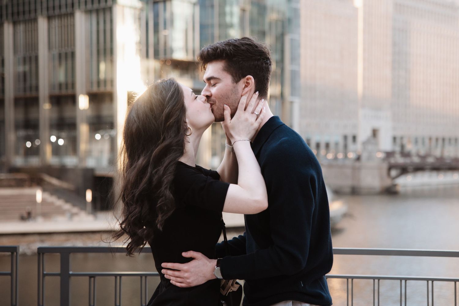 Romantic couple sharing a kiss on a bridge with a city skyline backdrop at golden hour.