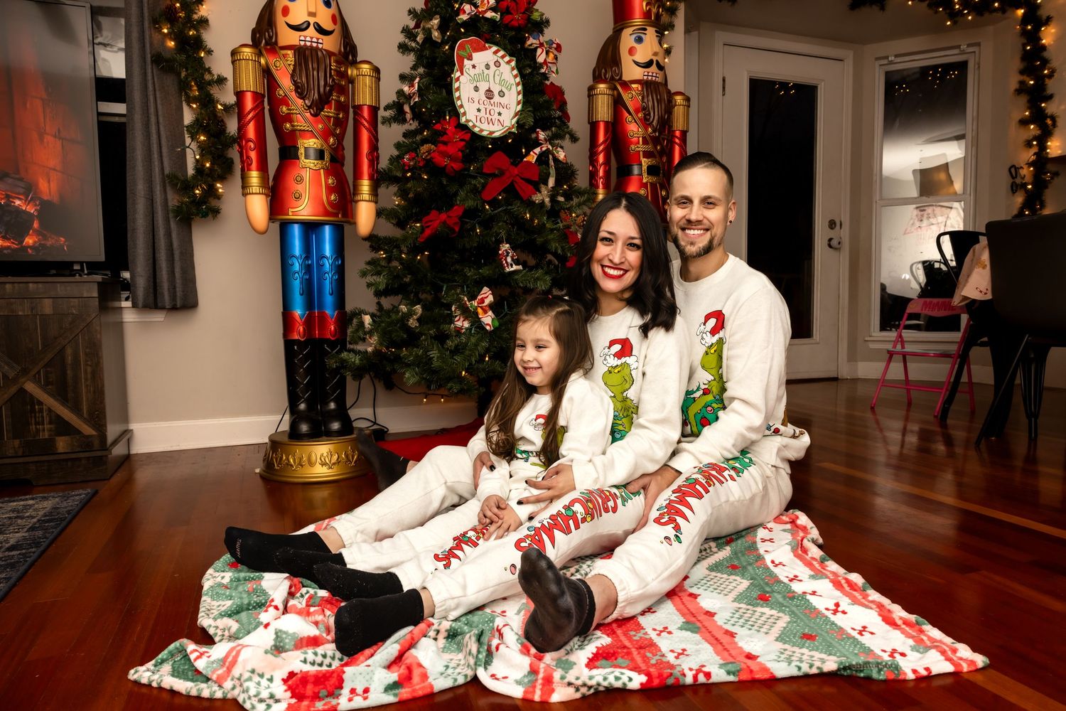 Smiling family of three in matching Grinch Christmas pajamas posing on a holiday blanket with nutcracker decor.