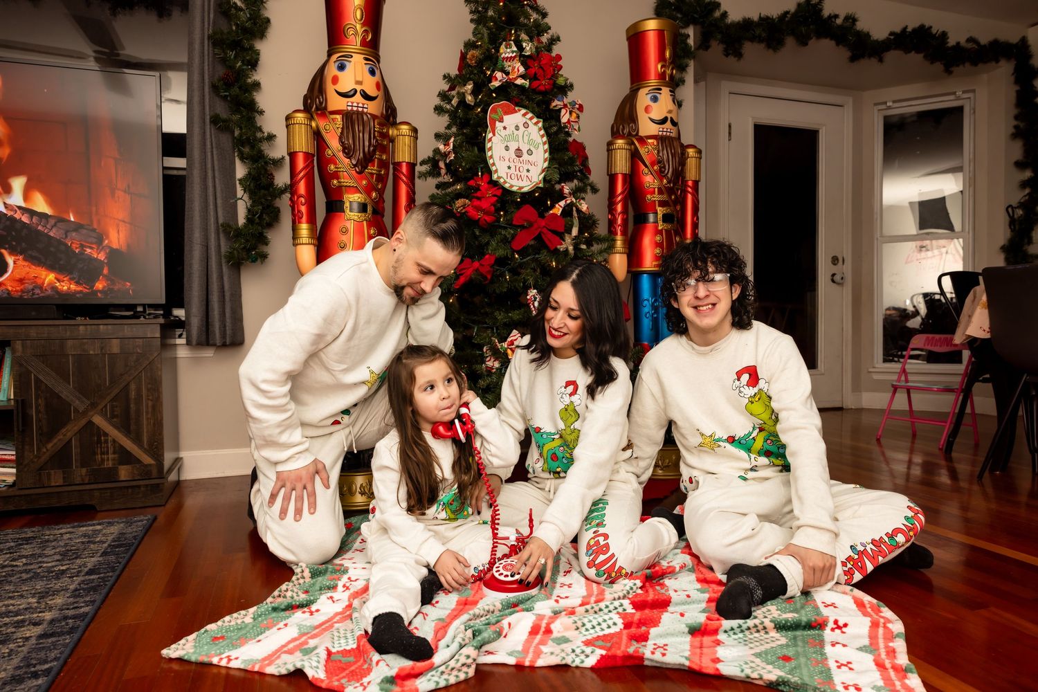 Family of four in matching Christmas pajamas sitting together on a holiday blanket near a decorated tree.