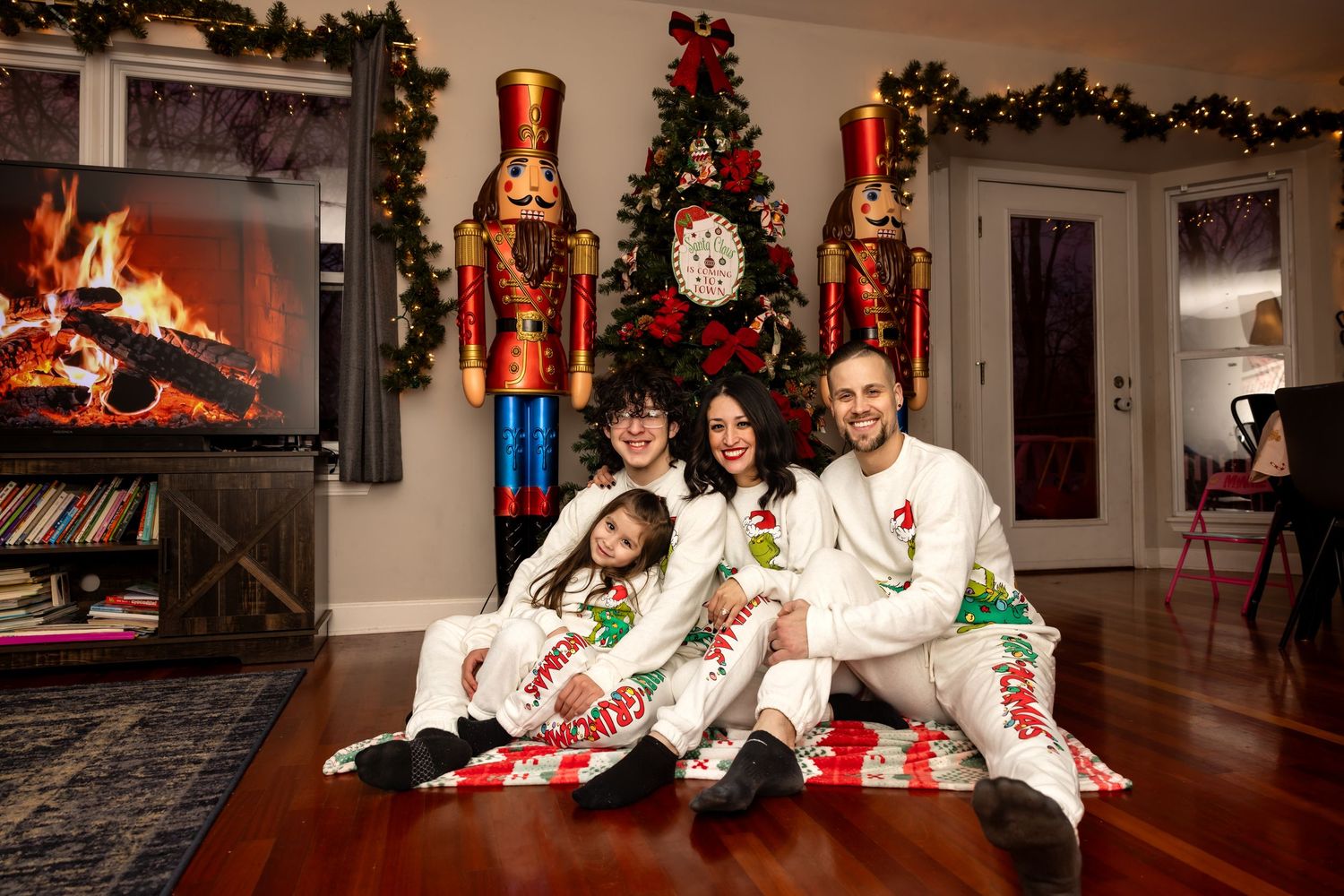 Happy family in matching white Grinch holiday pajamas sitting together by a Christmas tree and fireplace.