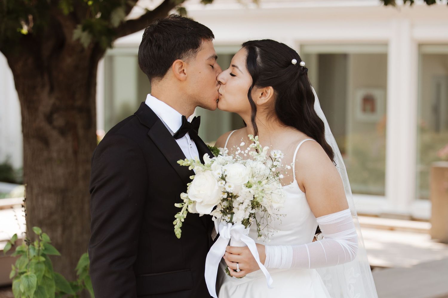 Bride and groom share a romantic kiss outdoors, bride holding white floral bouquet in wedding attire.