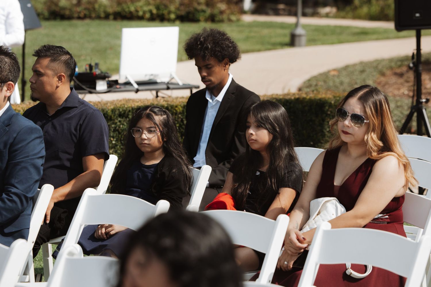 Wedding guests including children seated in white chairs watching outdoor garden ceremony.