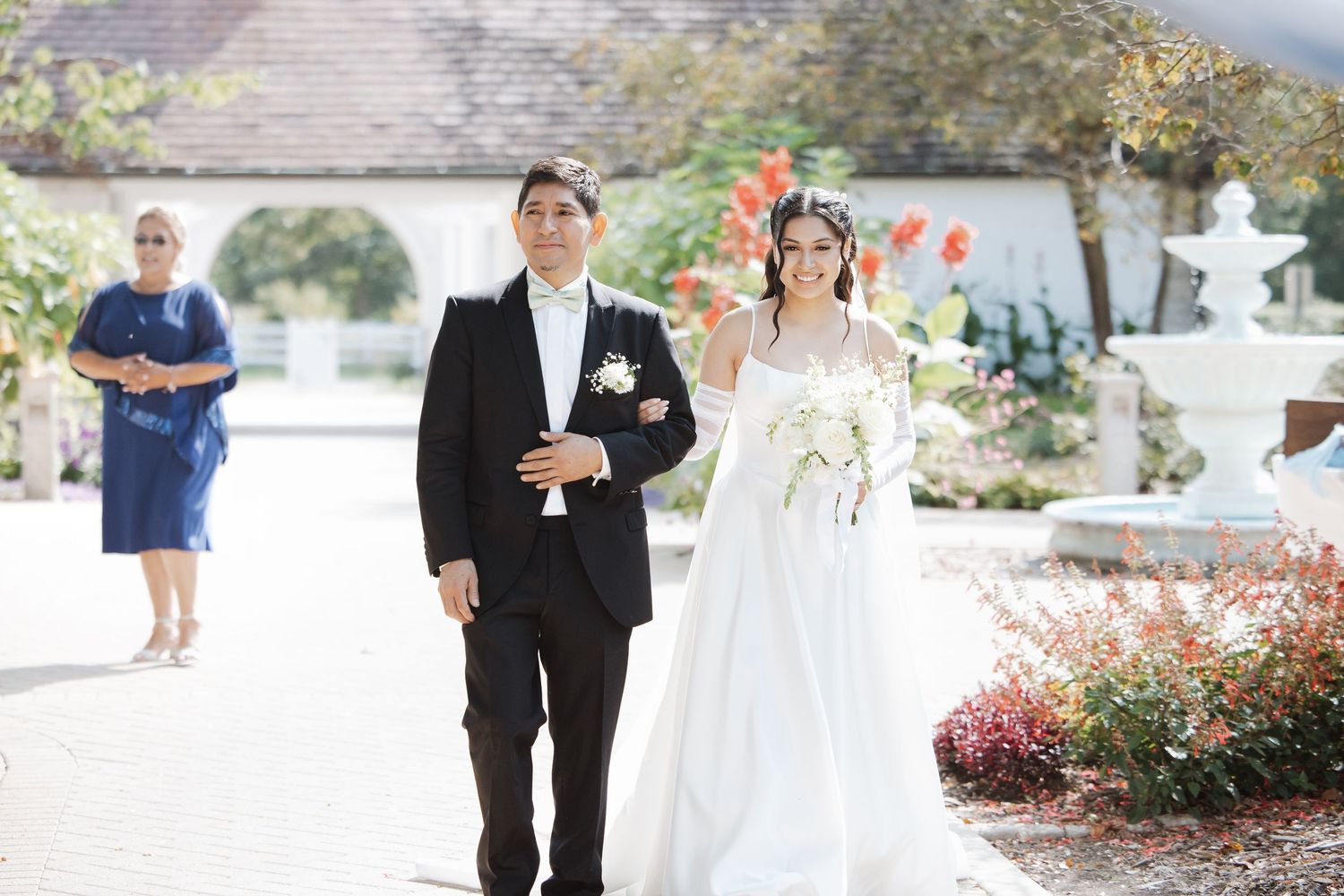 Bride in white gown walks down garden aisle escorted by man in black tuxedo.