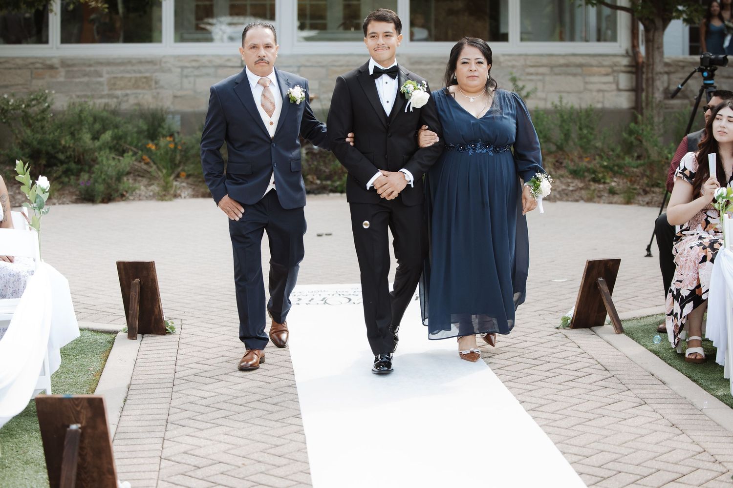Groom in black tuxedo walks down aisle escorted by parents at outdoor wedding ceremony.