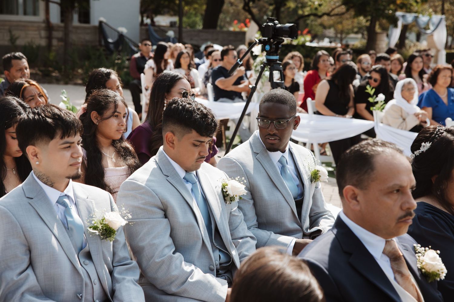 Groomsmen in gray suits seated among wedding guests during outdoor daytime ceremony.