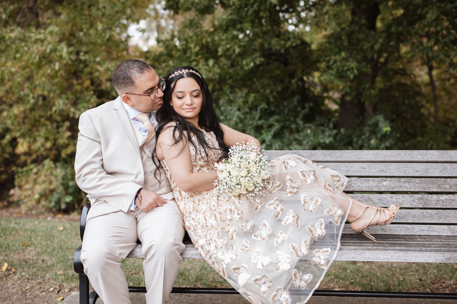 Groom tenderly looking at bride seated on park bench in gold lace wedding dress.