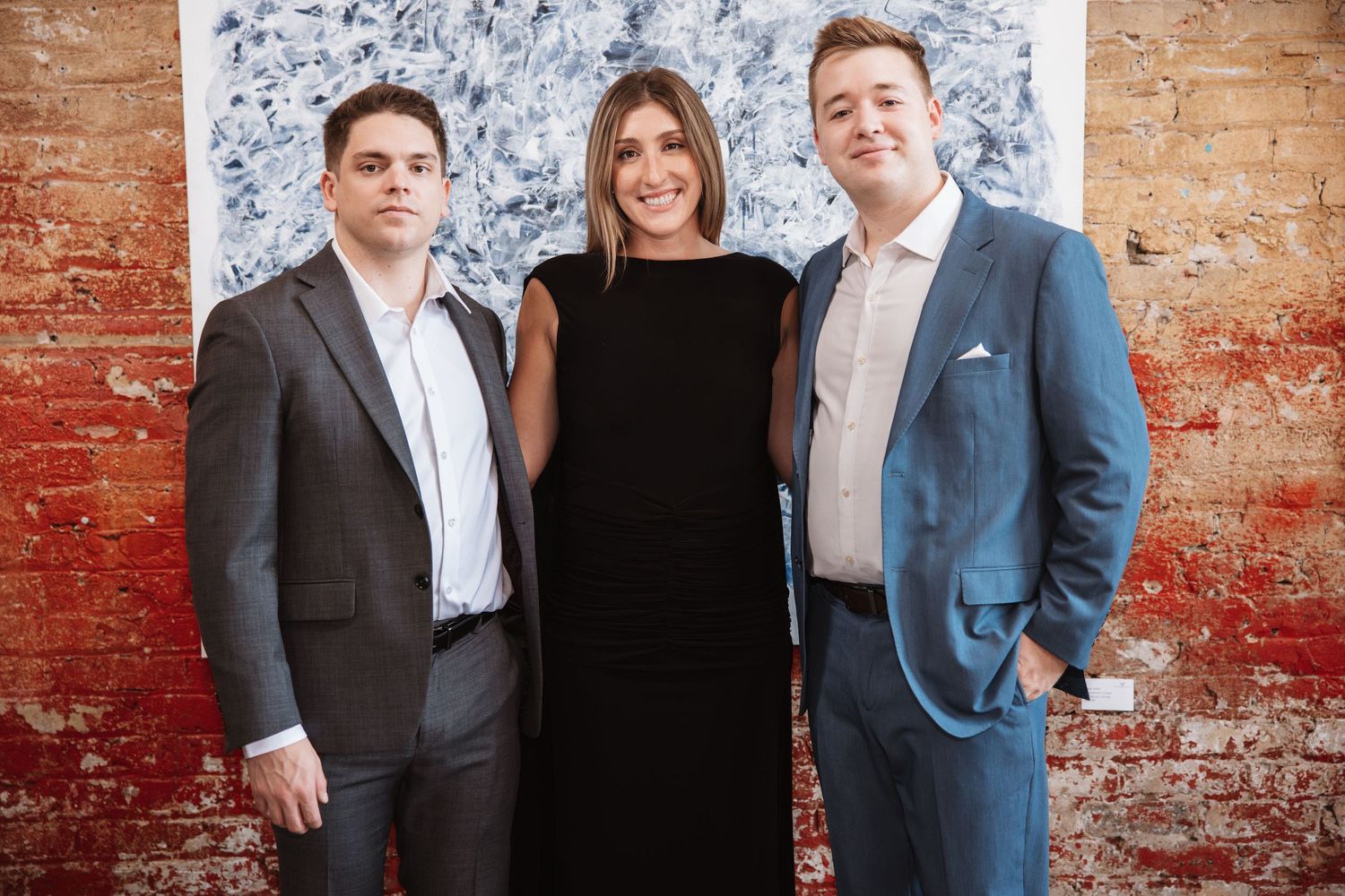 Three elegantly dressed professionals pose together at an art gallery event in front of abstract artwork.
