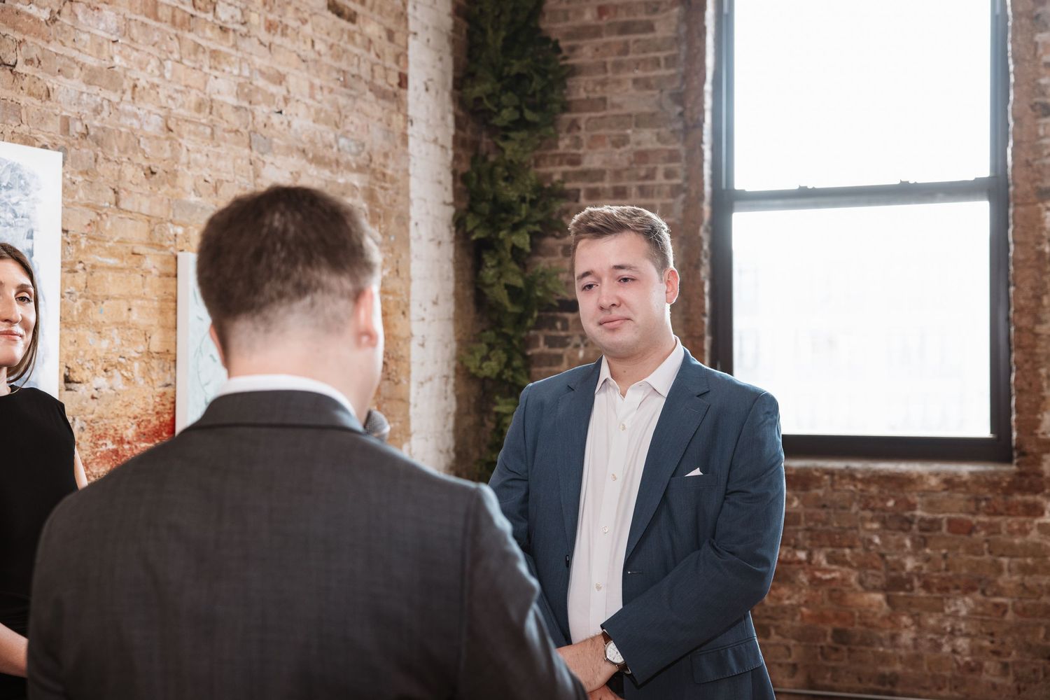 Two men in suits shaking hands in a rustic brick-walled venue with natural window light, color photo.
