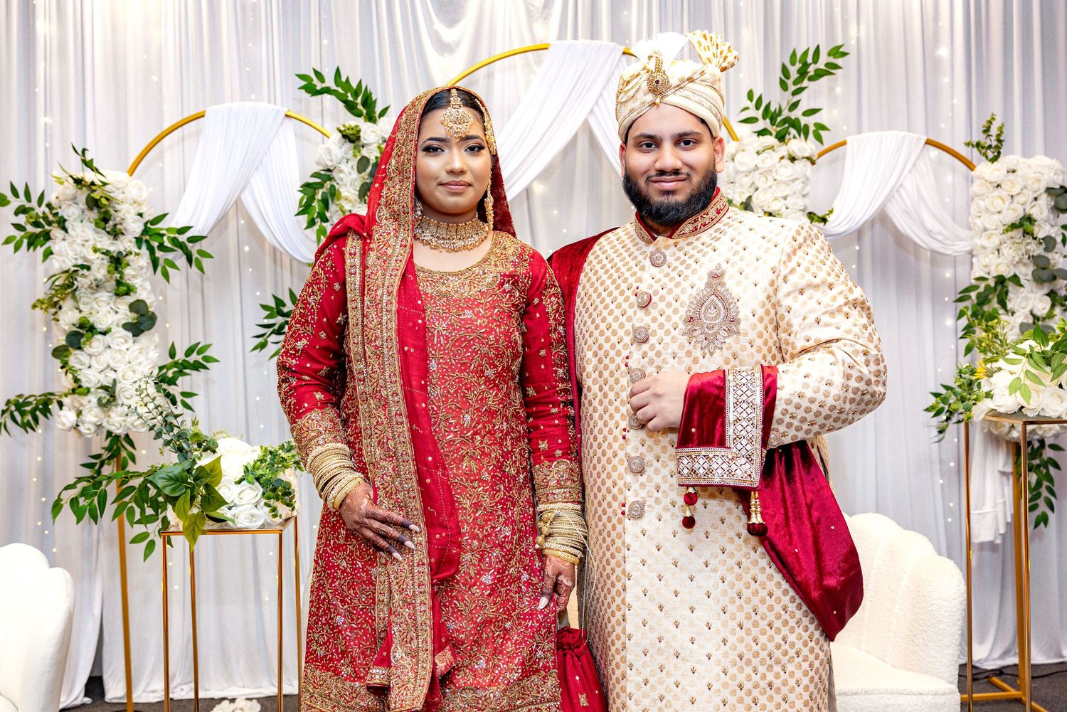 South Asian bride and groom posing in traditional red and gold wedding attire before white floral backdrop.