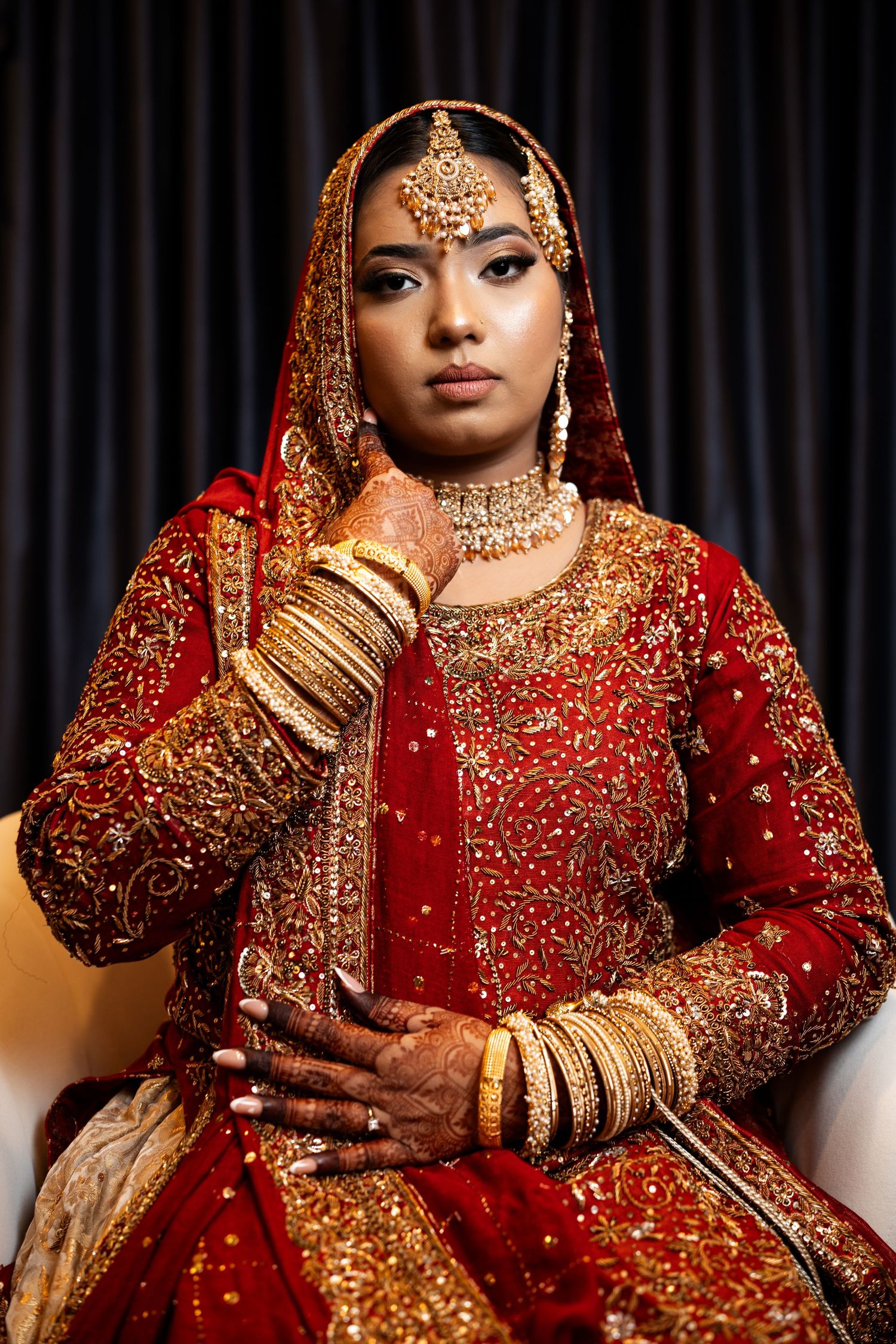 An Indian bride in a richly embroidered red and gold lehenga poses gracefully wearing traditional gold jewelry.