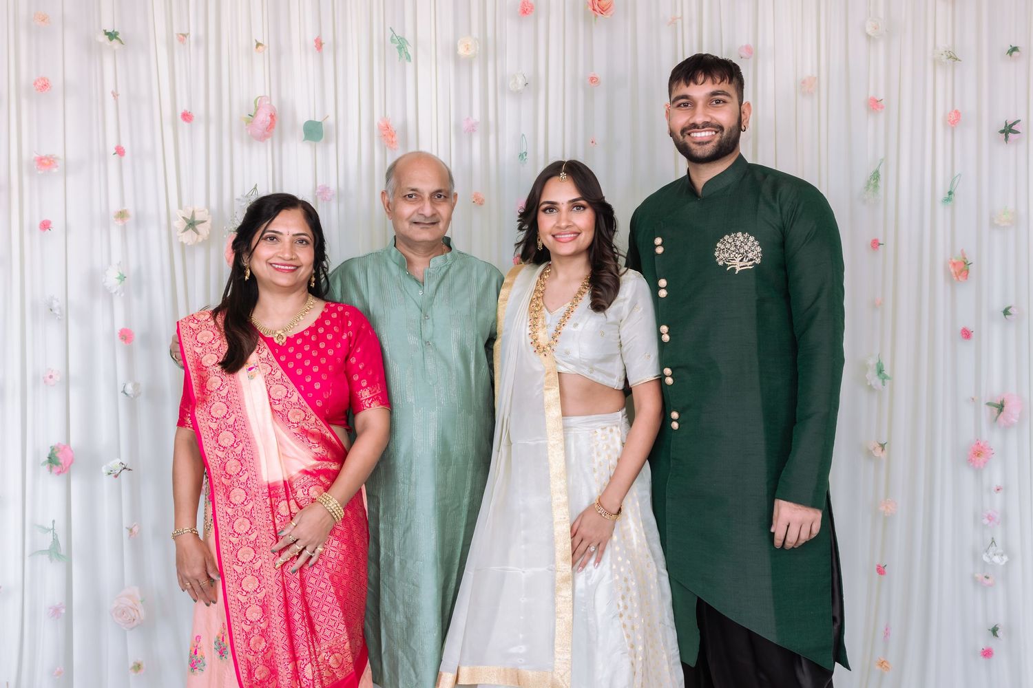 Indian family of four posing together at engagement ceremony with floral white backdrop.