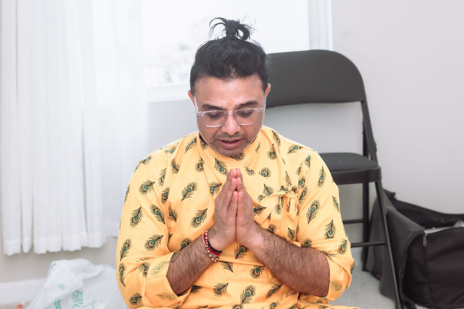 Man in yellow kurta with hair bun offering namaste greeting during Indian wedding celebration.