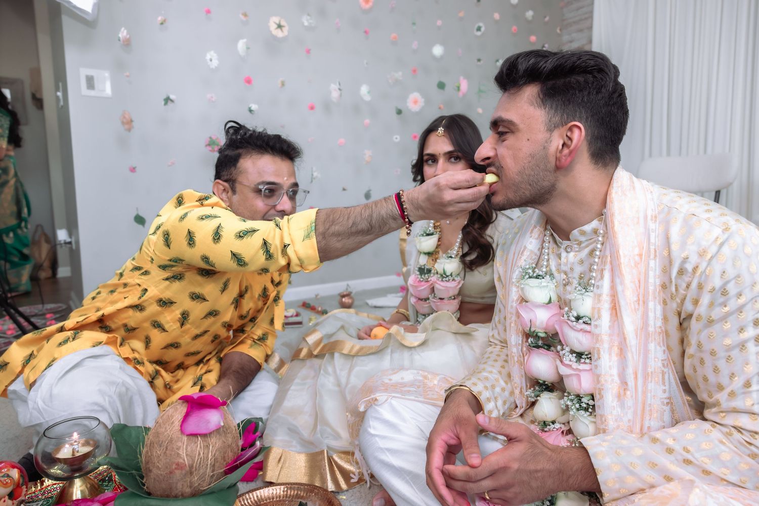 Hindu wedding ritual with man in yellow kurta offering food to groom in white sherwani during ceremony.