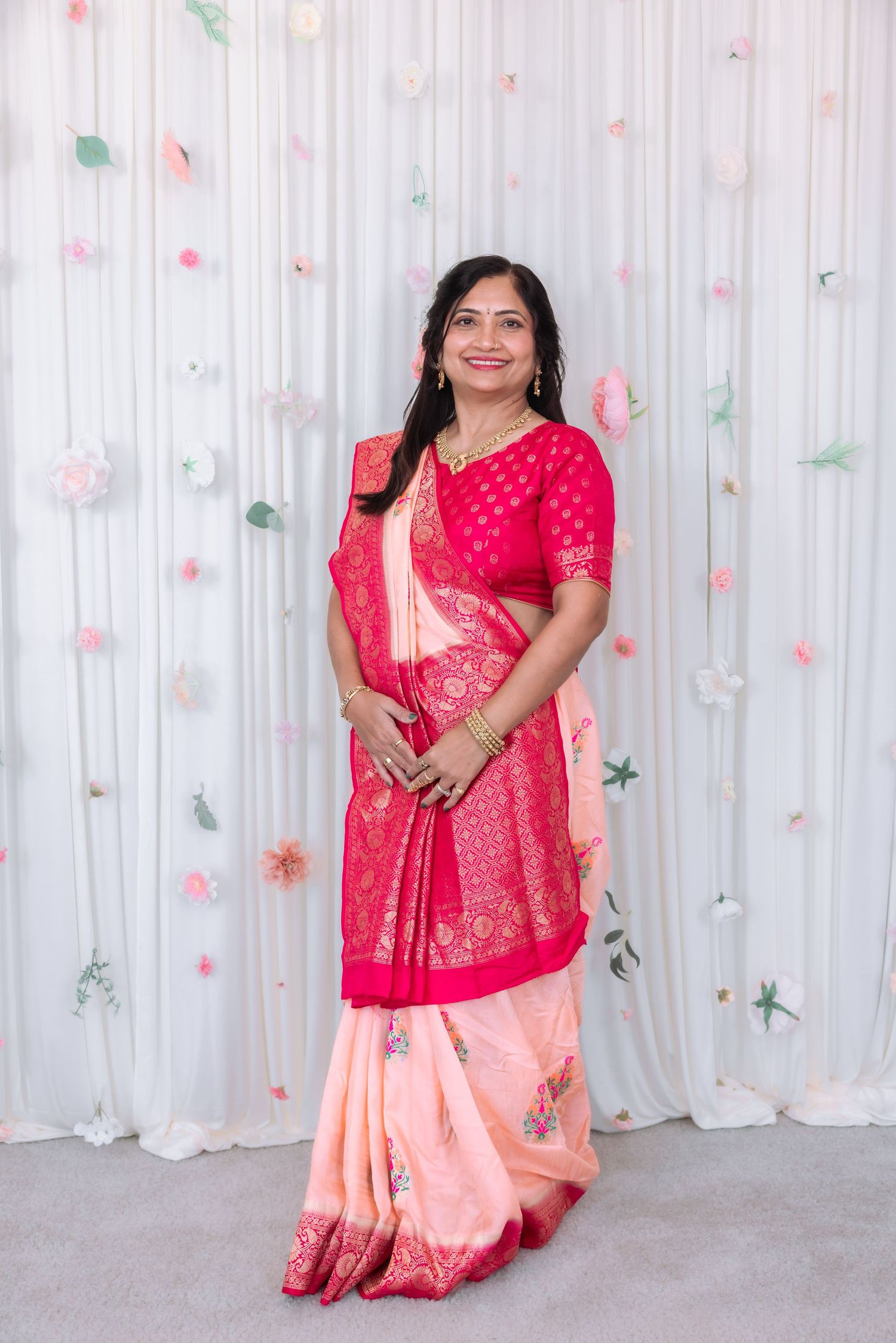 Elegant Indian woman in pink and red Banarasi saree poses confidently at floral decorated white wedding backdrop.