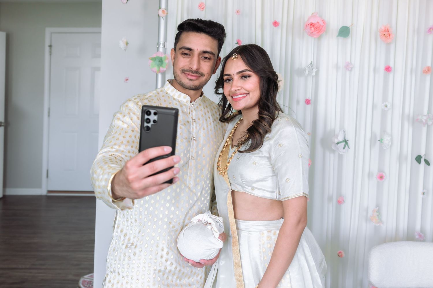 Young South Asian couple in traditional Indian attire taking selfie at decorated celebration event.