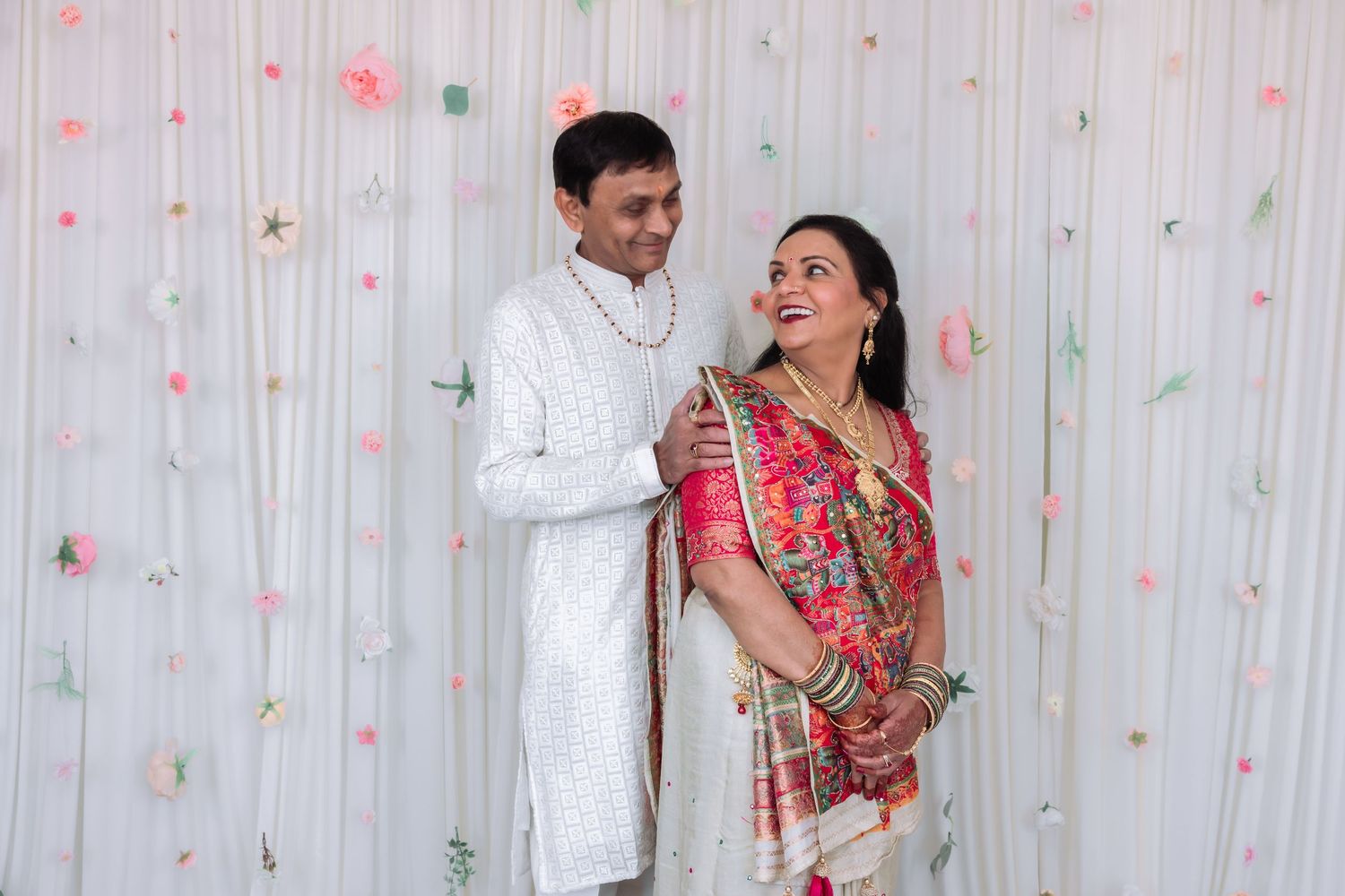 Indian couple in traditional white and pink attire sharing a joyful moment at a festive celebration.