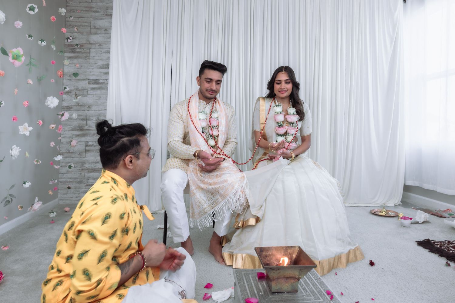 Bride and groom smile during a traditional Indian wedding ceremony with a seated priest.