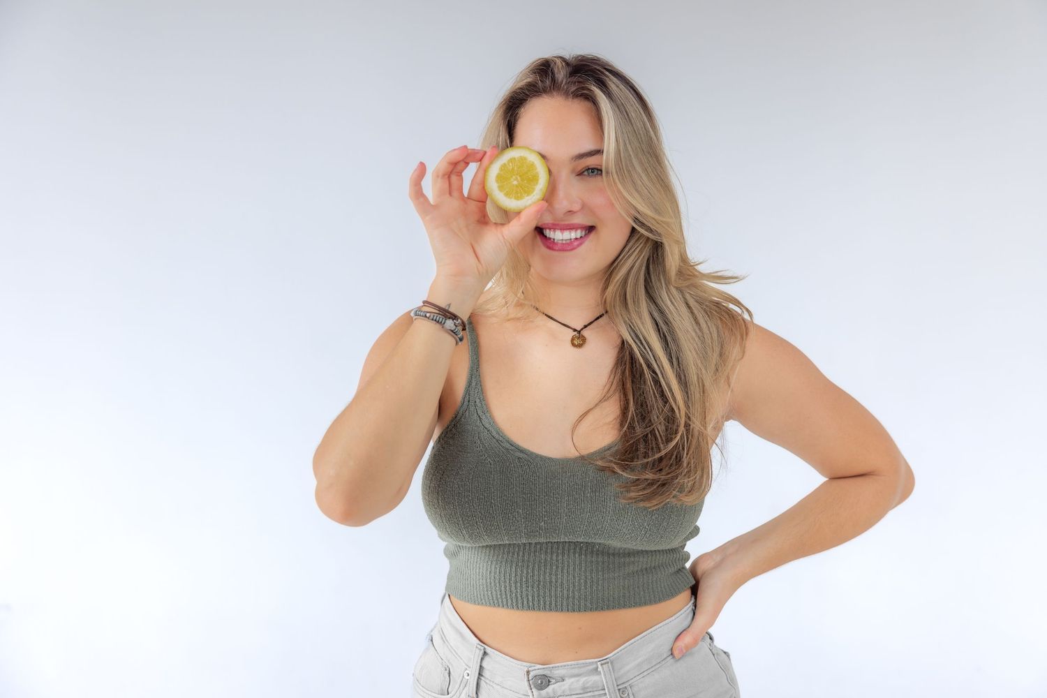Smiling blonde woman in green crop top holding lemon slice over eye against white background.