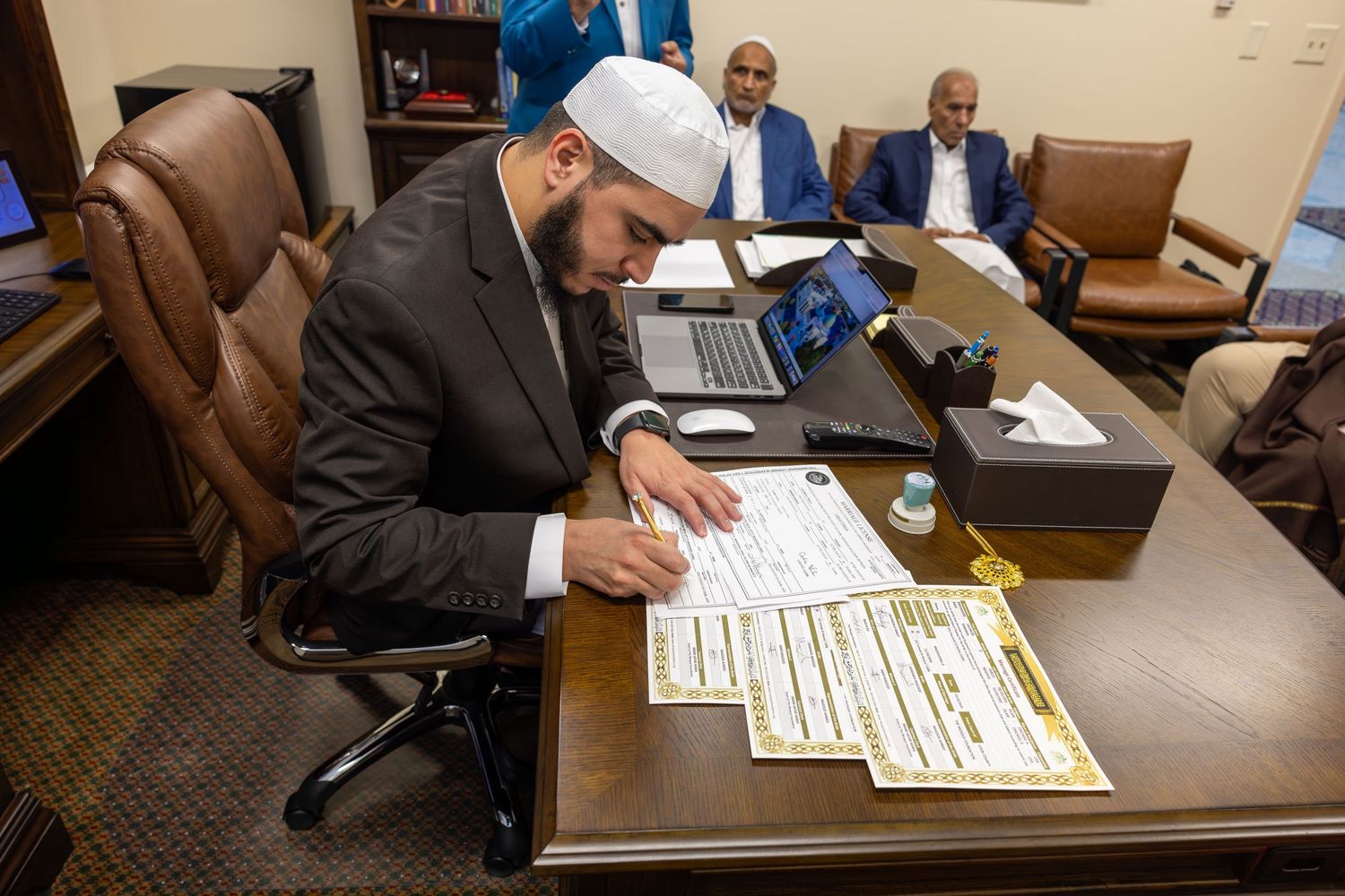 Imam in white kufi signing official Islamic Nikkah marriage certificate documents at an office desk.