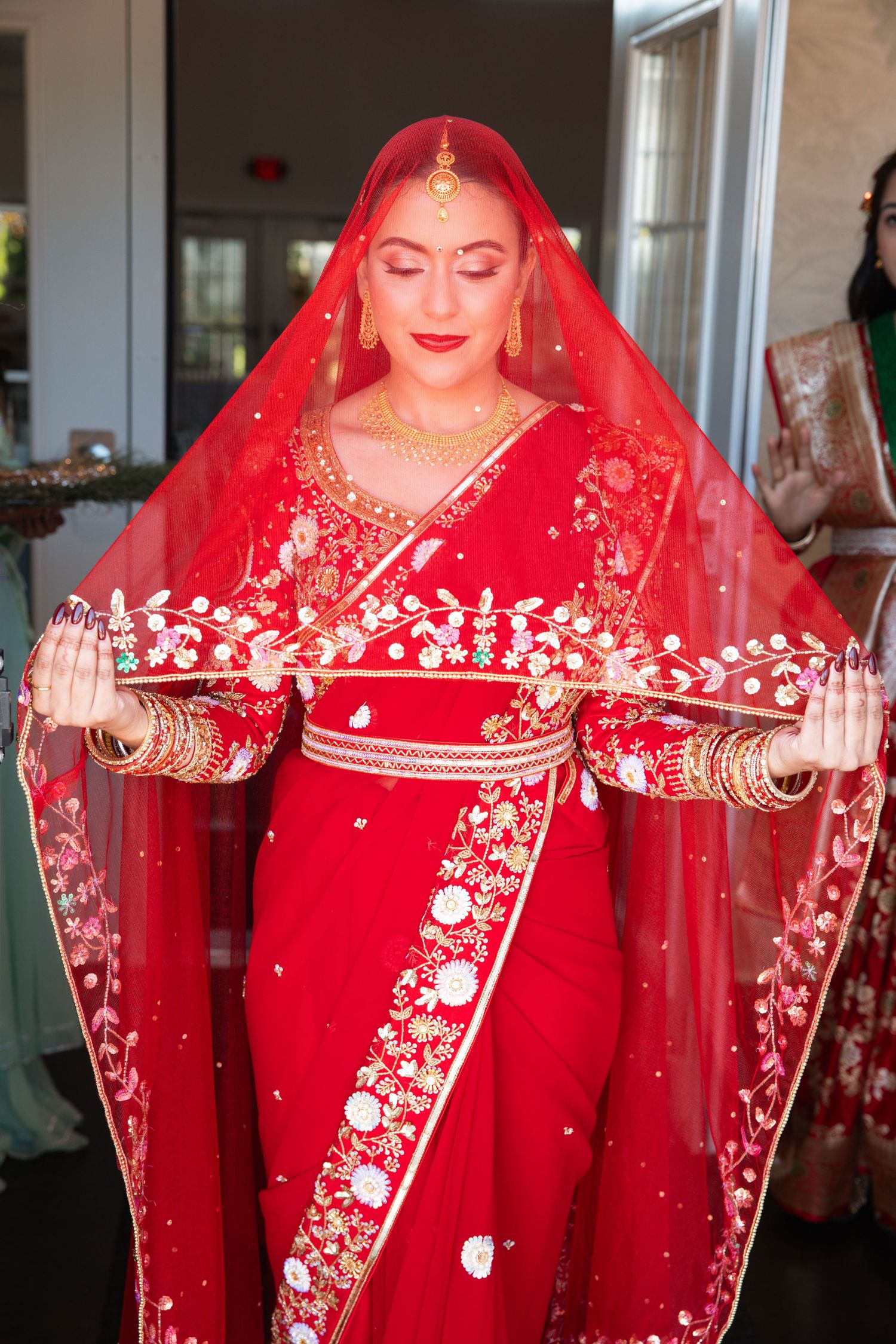 Indian bride in ornate red and gold bridal lehenga with embroidered dupatta and traditional gold jewelry at wedding ceremony.