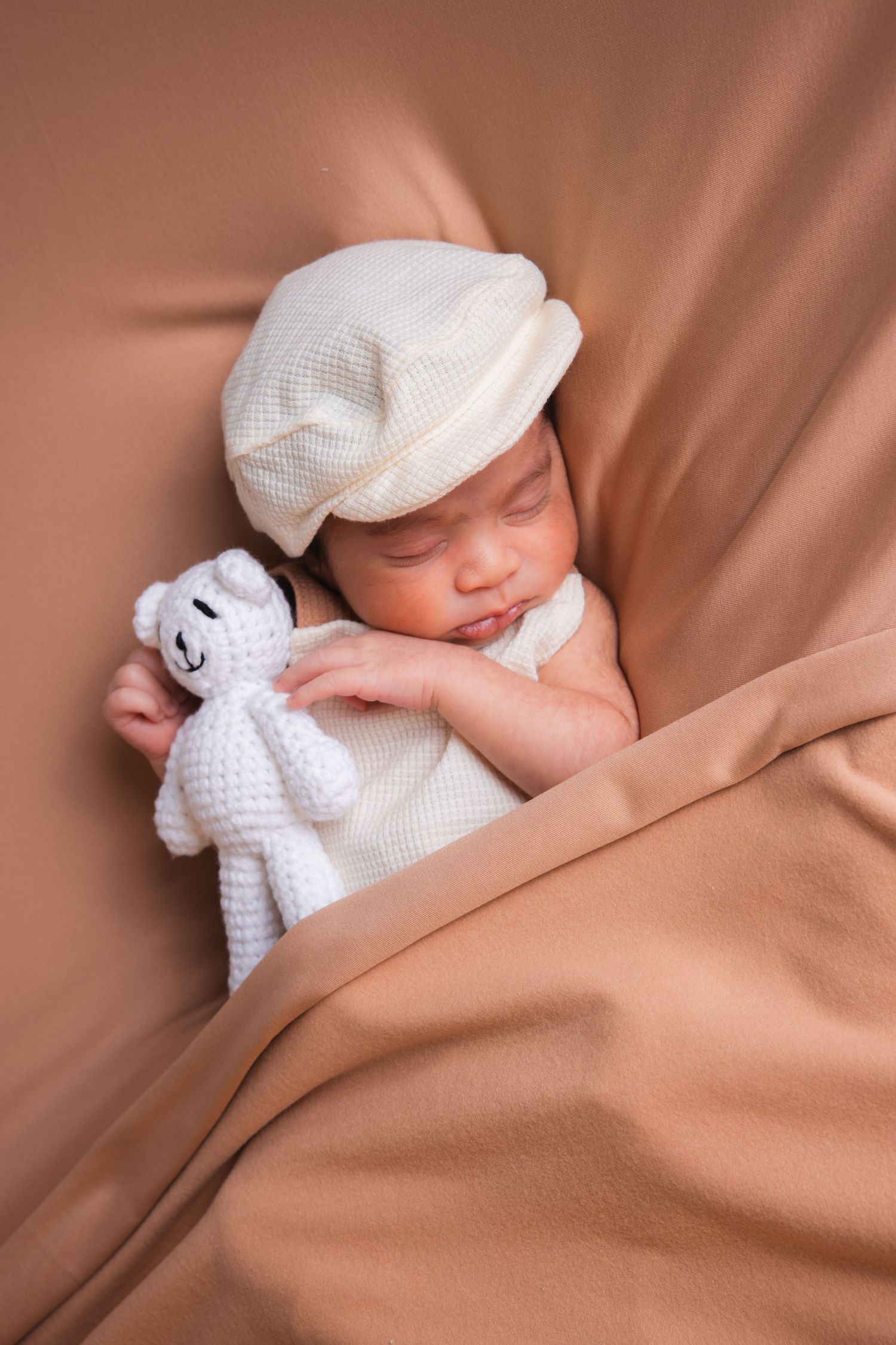 Peaceful newborn baby in white cap sleeps while holding a crocheted white teddy bear.