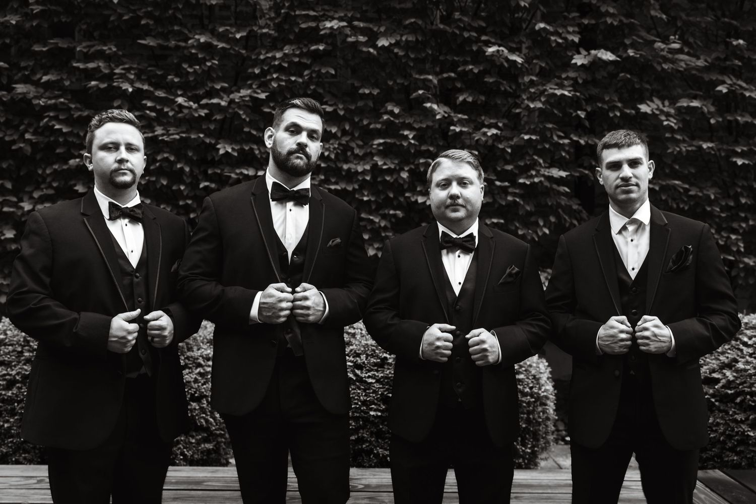Black and white photo of four groomsmen in tuxedos adjusting their lapels outdoors.