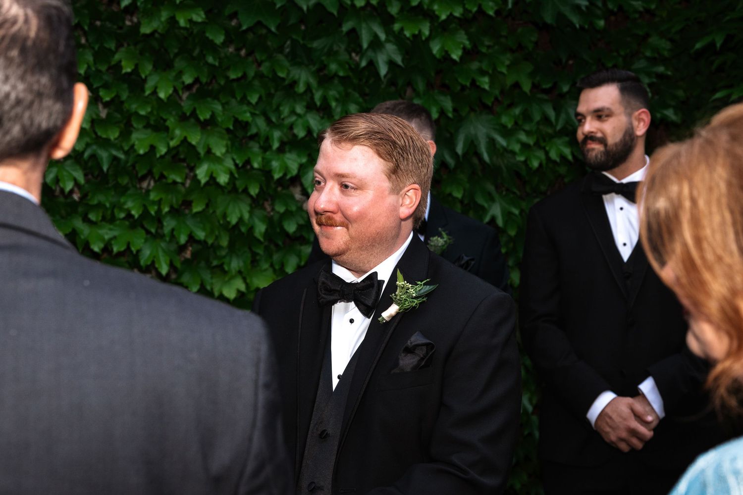 Happy groom in black tuxedo smiling during outdoor wedding ceremony with lush green ivy wall behind him.