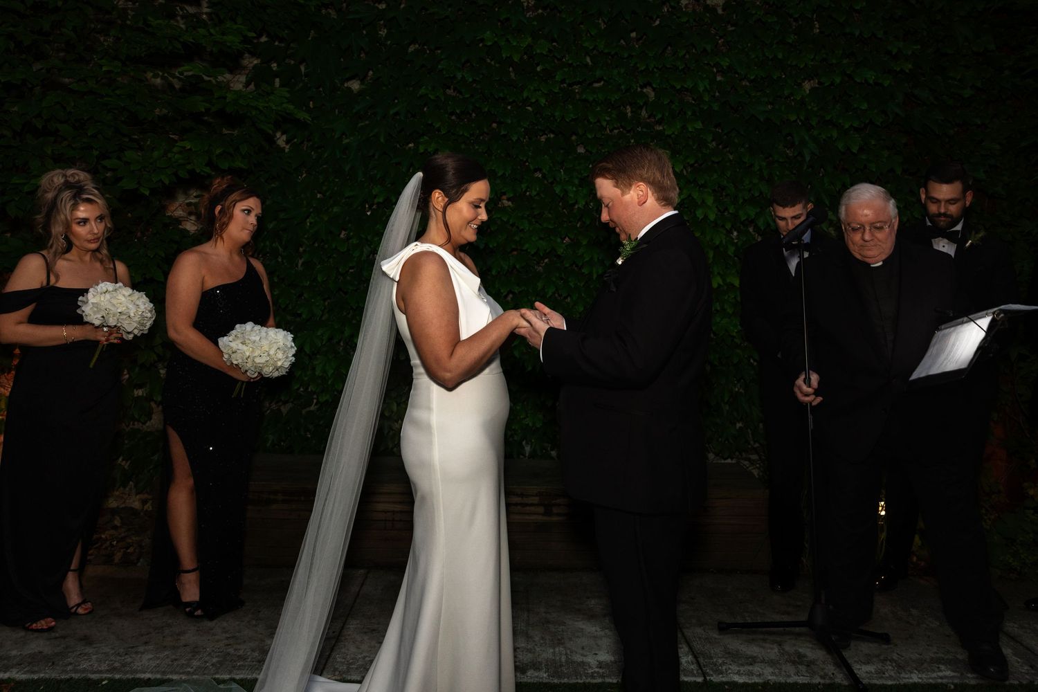 Bride and groom exchange rings at outdoor wedding ceremony with bridesmaids in black dresses.