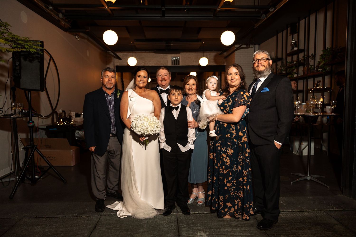 Bride and groom pose with seven family members including young children at warmly lit wedding reception.