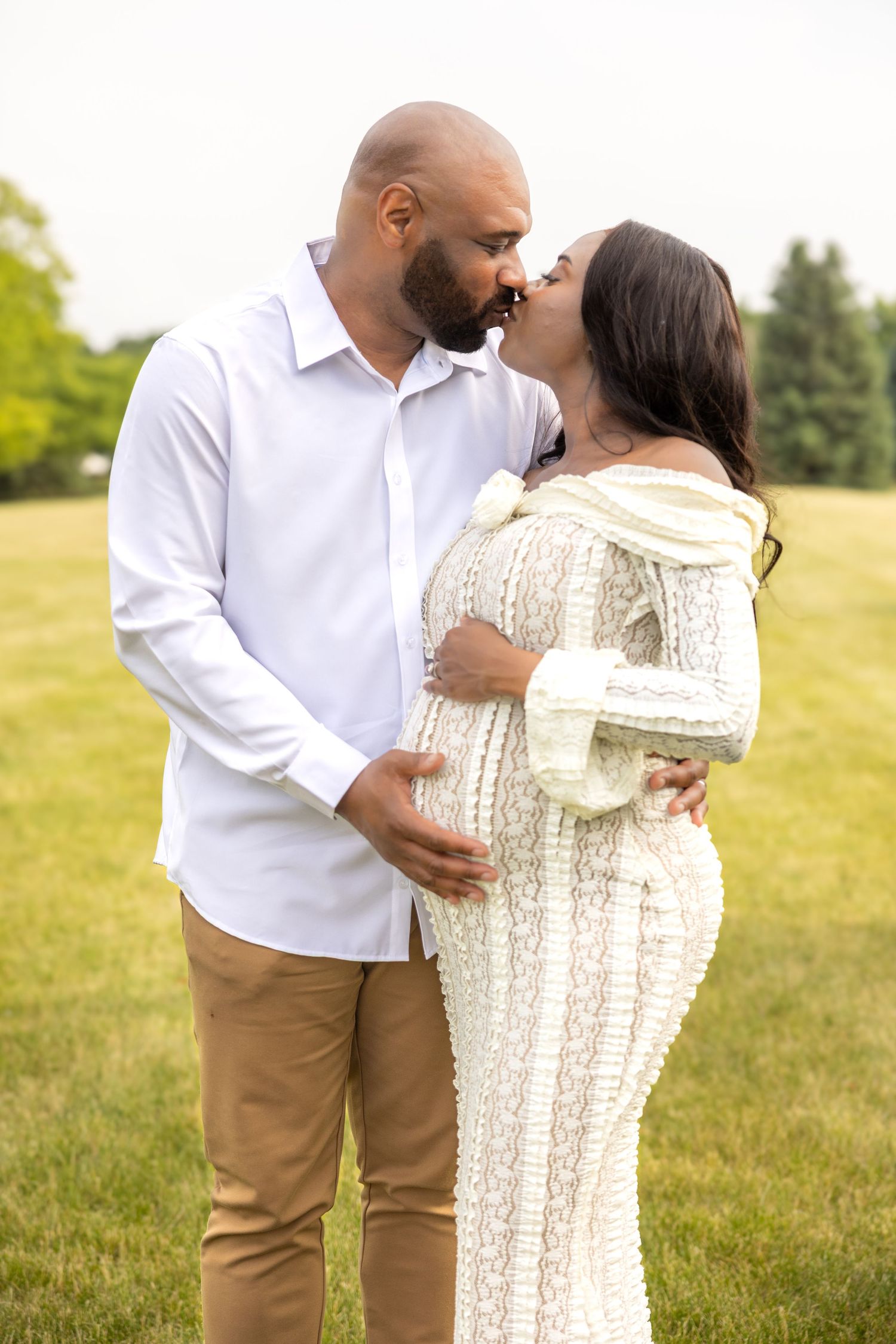 Couple sharing a kiss outdoors during maternity photoshoot, woman in white lace gown.