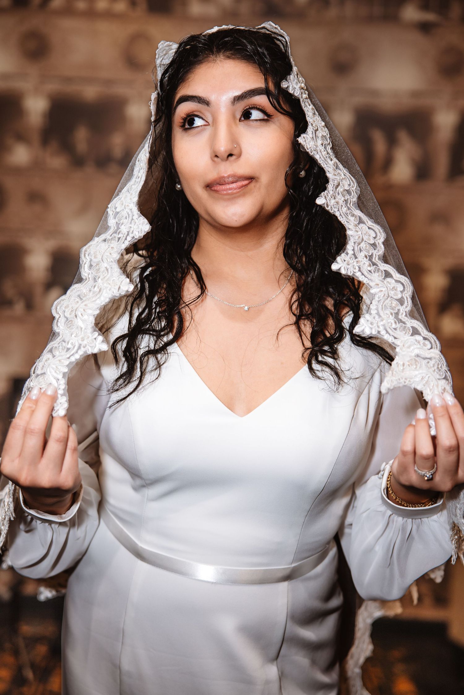 Beautiful bride in white satin dress and lace veil poses with raised hands.