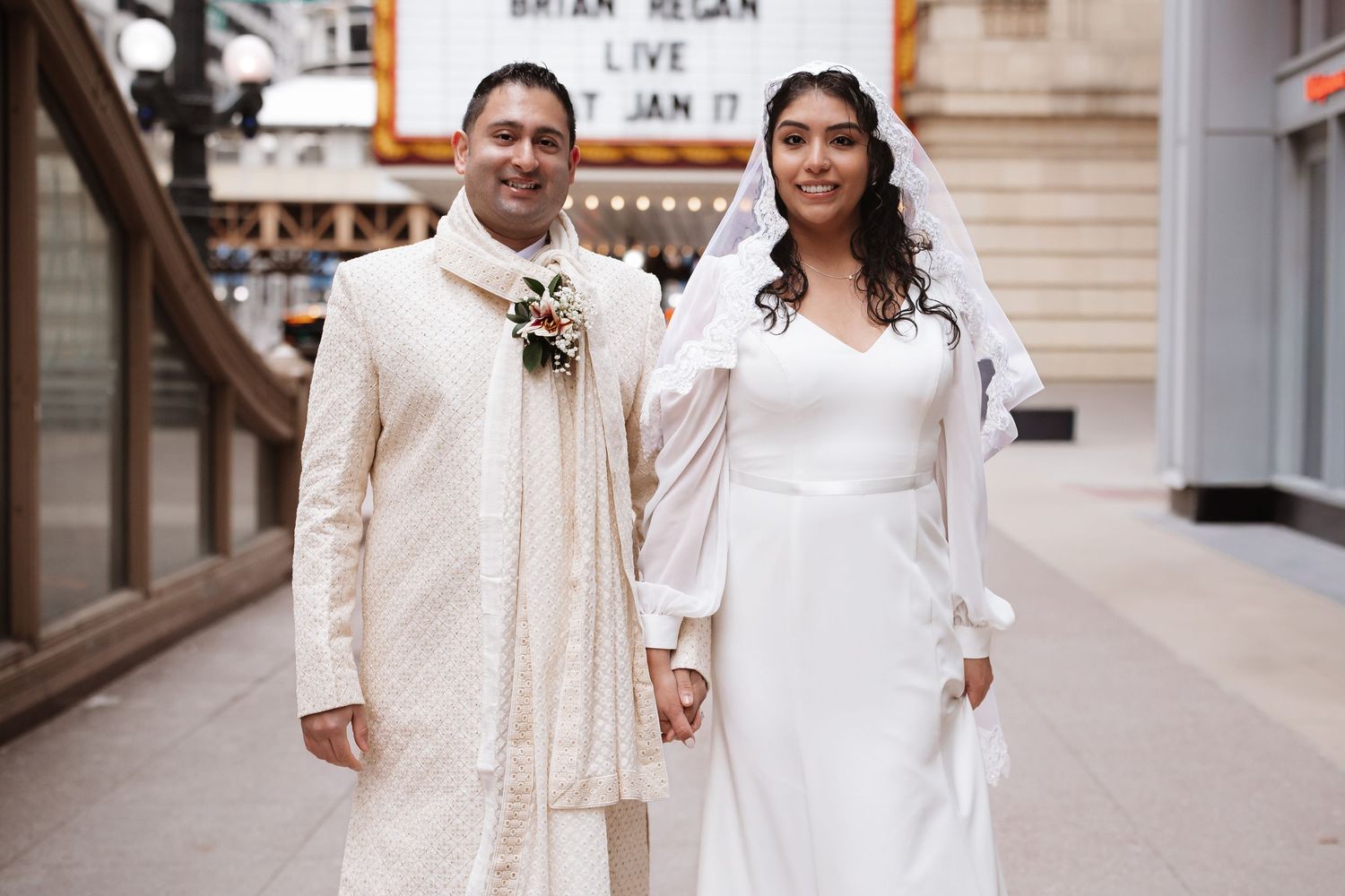 Indian groom in cream sherwani and bride in white gown pose together near Chicago Theatre.