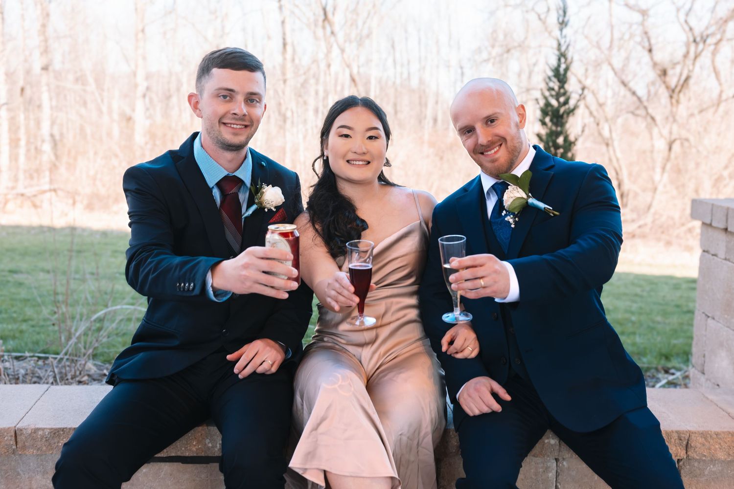 Two groomsmen in navy suits and a woman in a champagne dress smiling and toasting at a wedding.