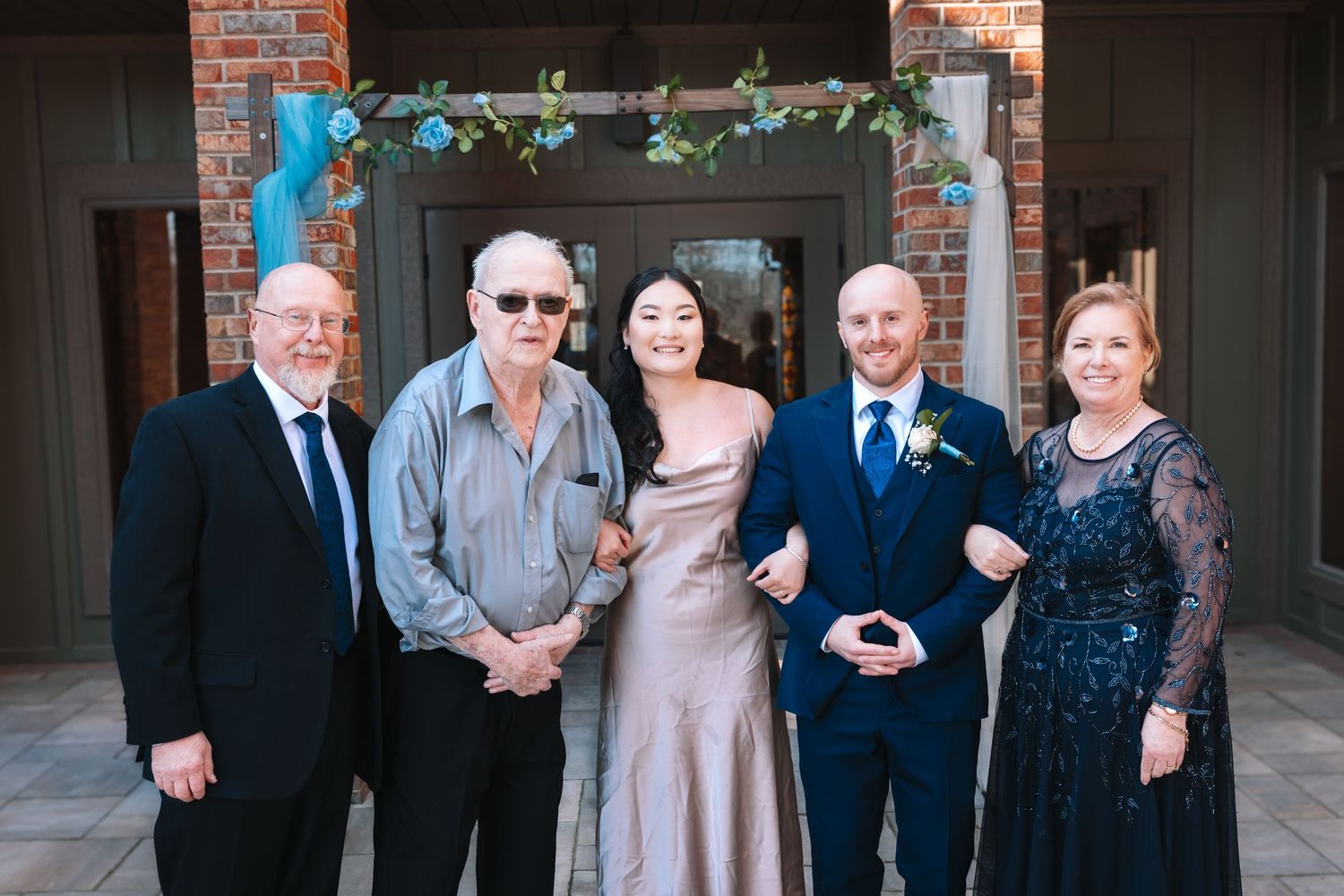 A bride and groom smile with three family members outside a brick venue adorned with blue floral decor.