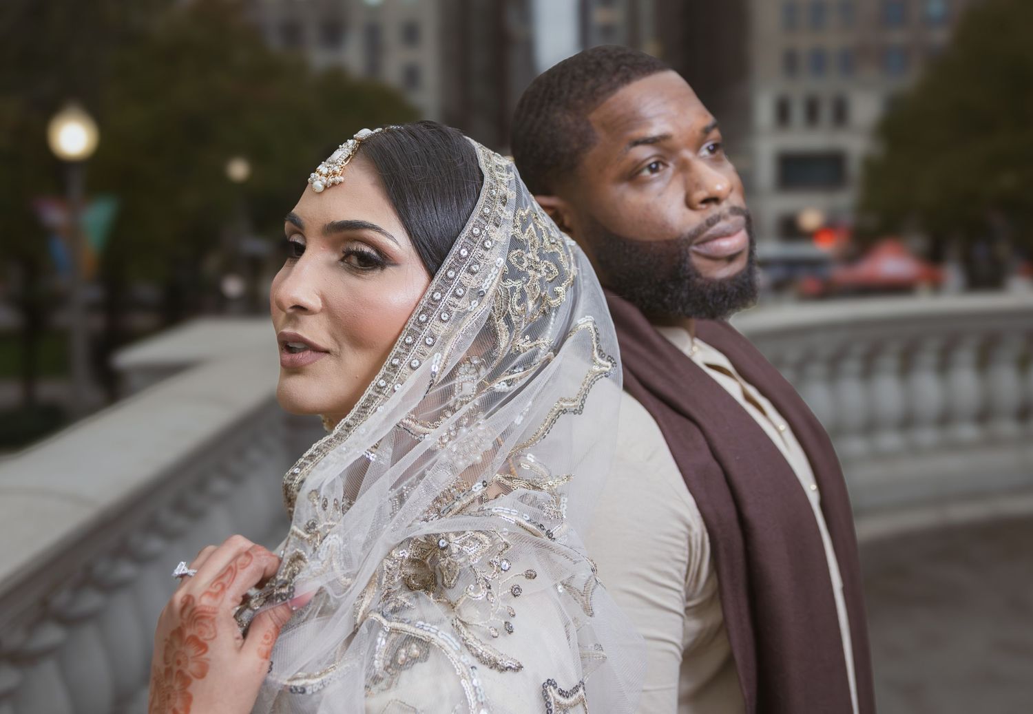 South Asian bride in embroidered hijab and groom in brown sherwani pose back-to-back in urban setting.