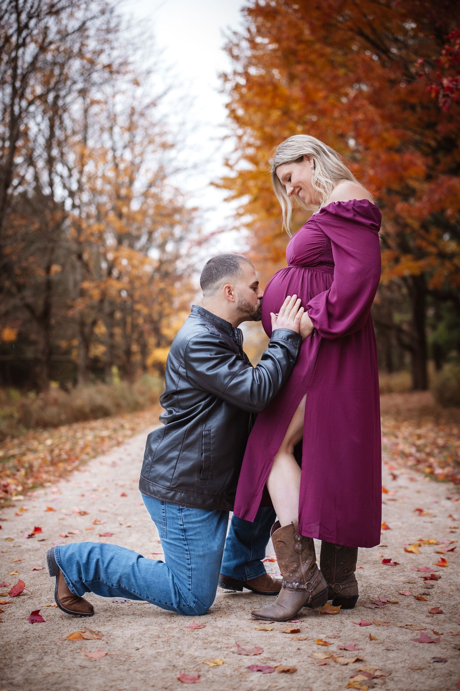 Man kneeling and kissing pregnant woman's bump as she smiles down, surrounded by fall foliage on a path.