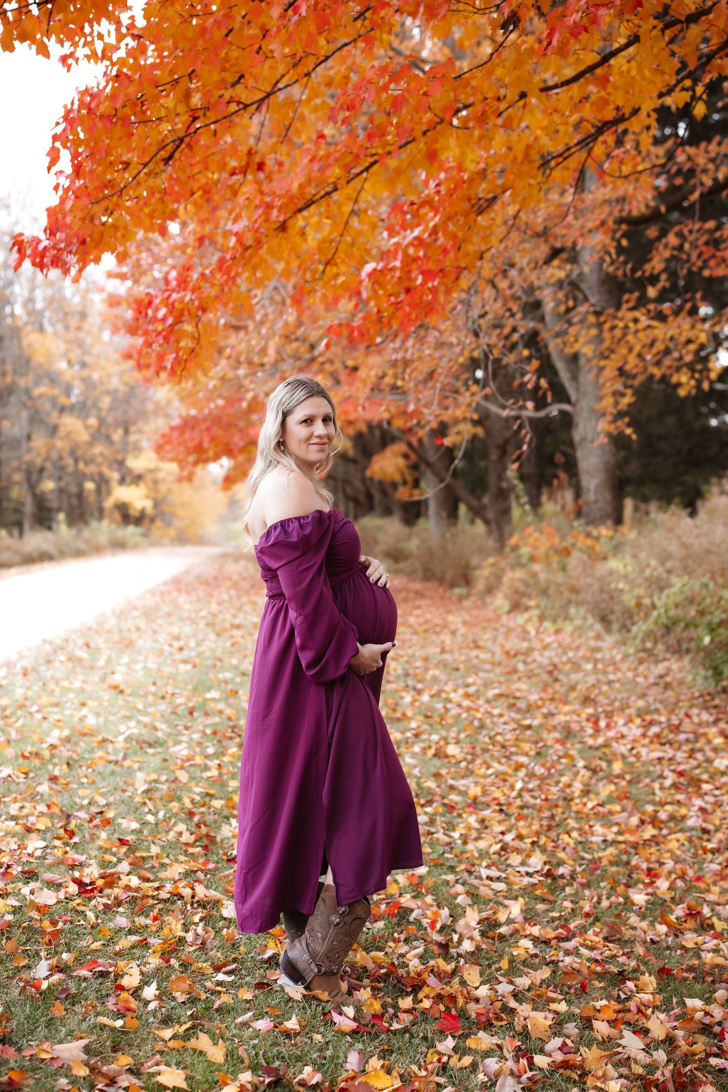 Pregnant woman in purple maternity dress posing on scenic autumn path lined with colorful trees.