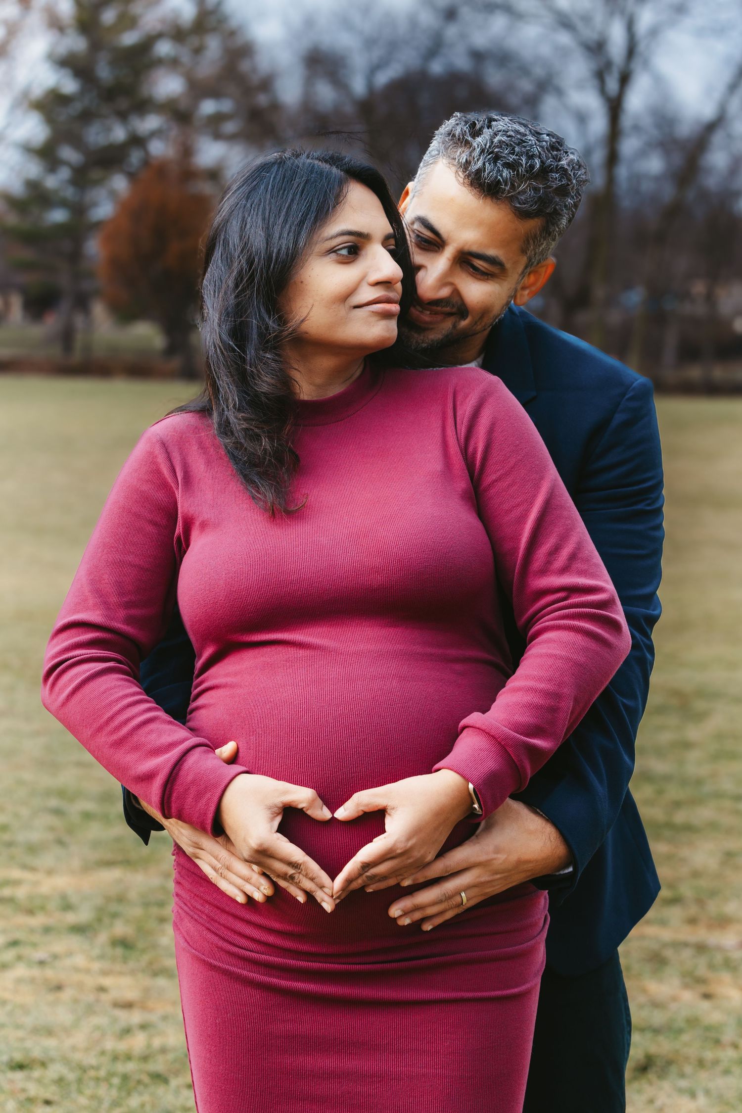 Pregnant couple in park, man in navy jacket kissing woman's cheek as both cradle her baby bump.