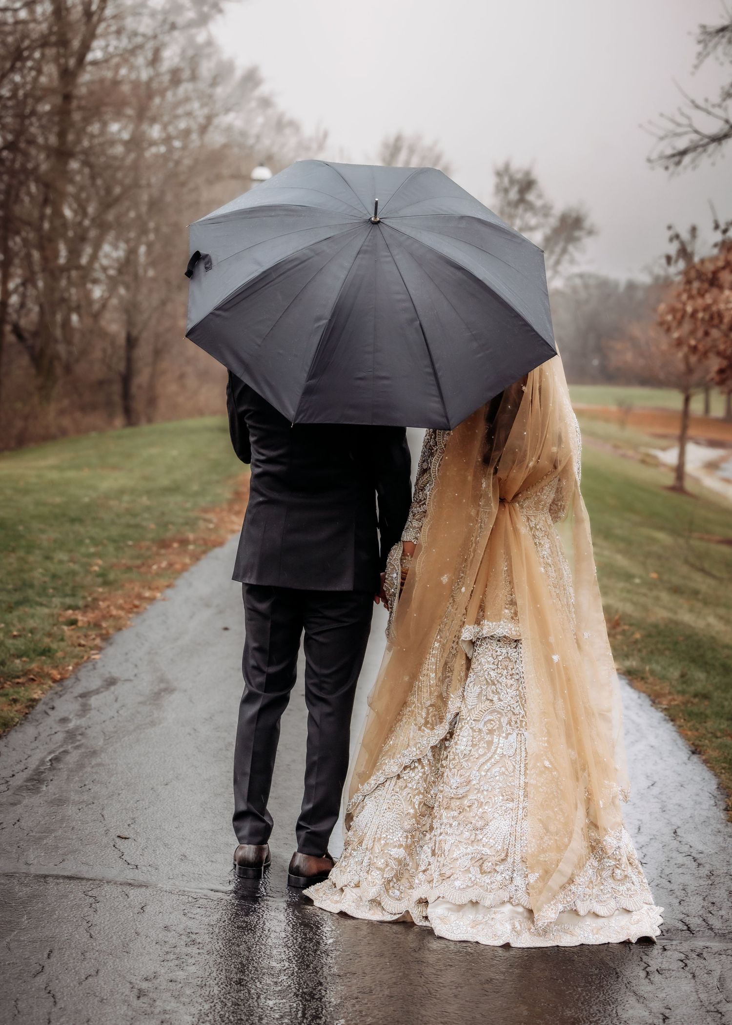 Bride and groom walking in rain under black umbrella, bride wearing gold embroidered lehenga.