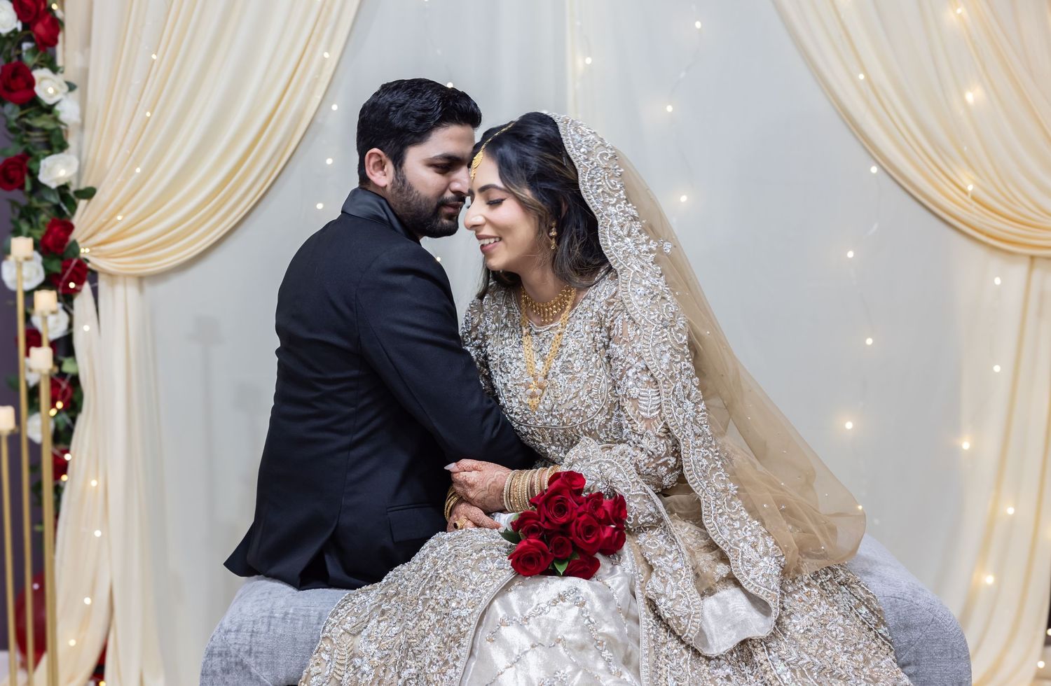 South Asian bride and groom share intimate moment holding red roses beneath decorated wedding backdrop.