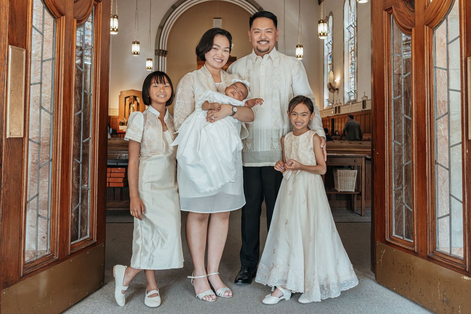 Filipino family in white formal attire poses at church entrance during baptism ceremony.