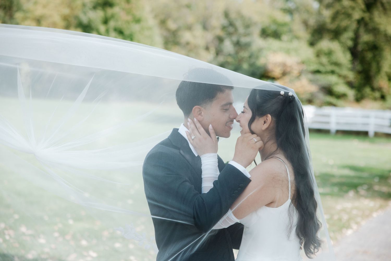 A bride and groom share a romantic moment under a flowing white veil outdoors on their wedding day.