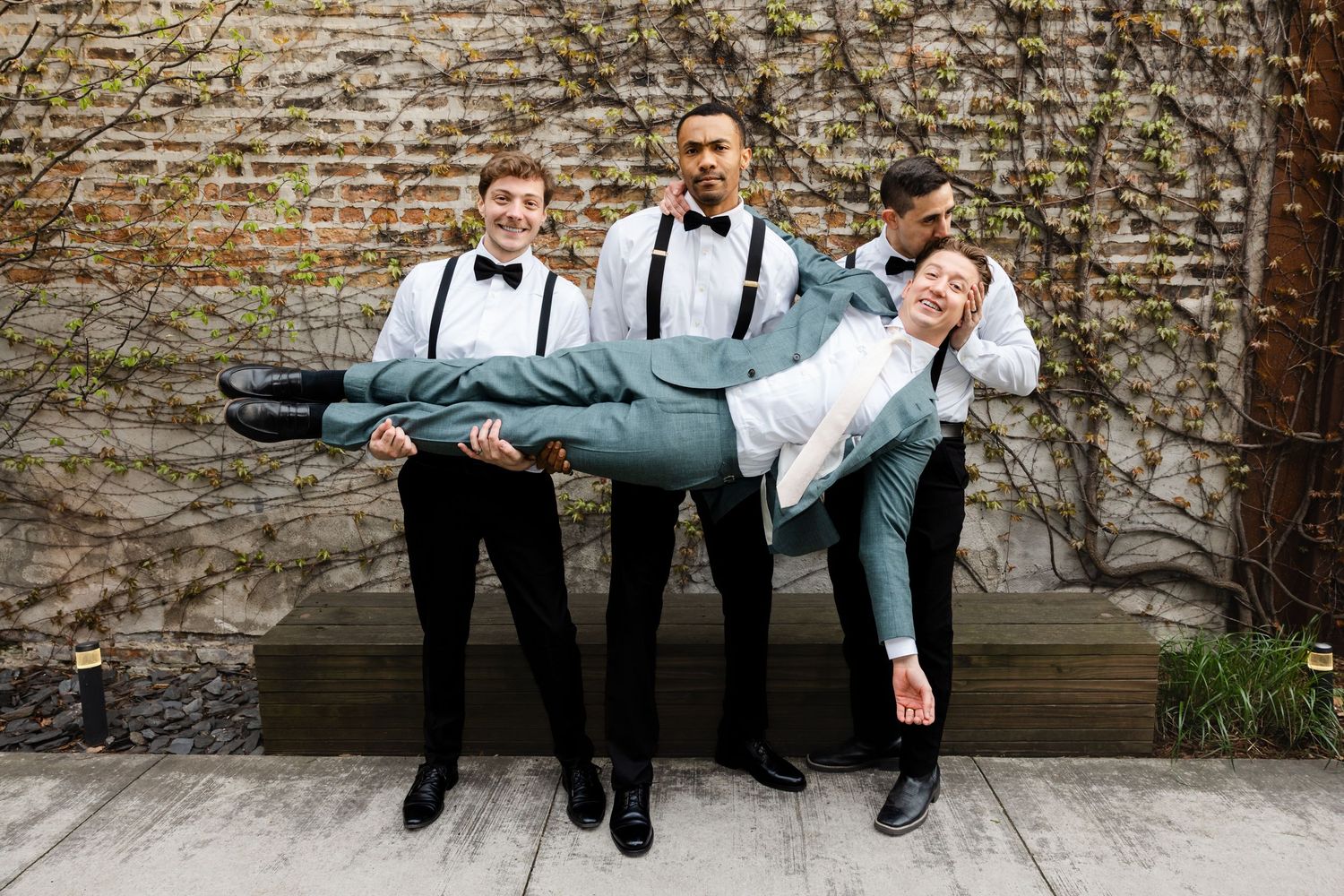 Three groomsmen in suspenders and bow ties playfully carry groom in teal suit outdoors near brick wall.